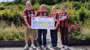 Four people standing side-by-side in a sunny garden setting, holding a large oversized AIB bank cheque. All four are wearing matching red, green, and white striped jerseys featuring the text "SULTANS OF PING". The cheque is made out to "Down Syndrome Cork Field of Dreams" for the amount of €10,163, signed by "FORAS".