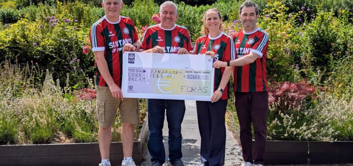 Four people standing side-by-side in a sunny garden setting, holding a large oversized AIB bank cheque. All four are wearing matching red, green, and white striped jerseys featuring the text "SULTANS OF PING". The cheque is made out to "Down Syndrome Cork Field of Dreams" for the amount of €10,163, signed by "FORAS".