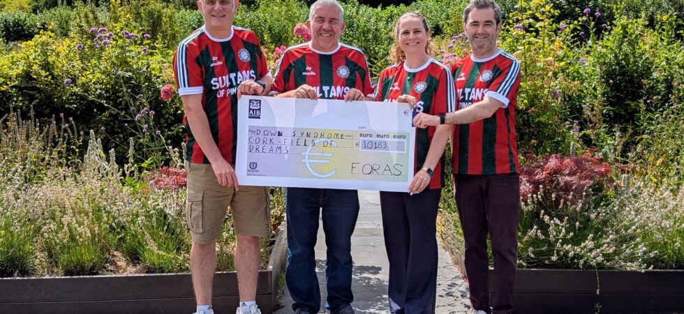 Four people standing side-by-side in a sunny garden setting, holding a large oversized AIB bank cheque. All four are wearing matching red, green, and white striped jerseys featuring the text "SULTANS OF PING". The cheque is made out to "Down Syndrome Cork Field of Dreams" for the amount of €10,163, signed by "FORAS".